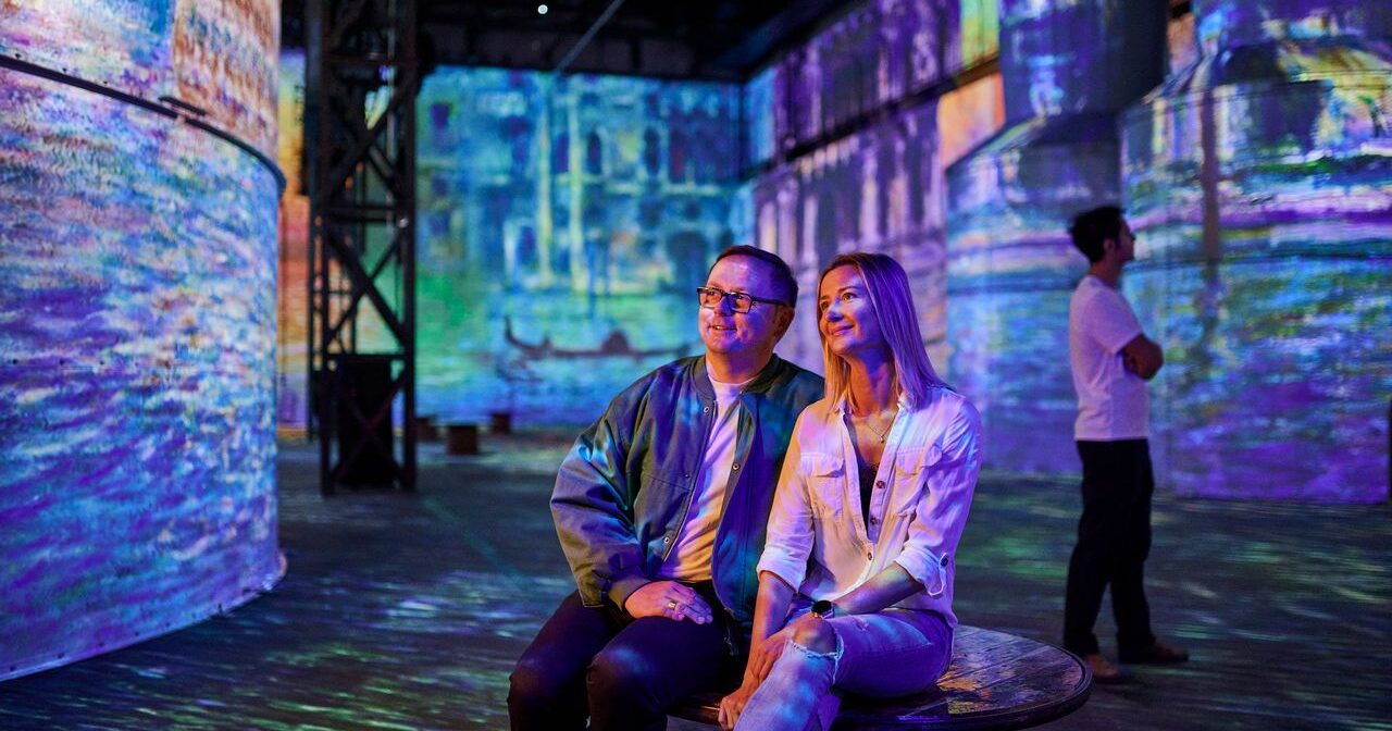A man and woman sit together on a bench, smiling and looking at colorful, immersive projections of artwork on the walls around them. Another person stands nearby, also observing the digital art exhibit.