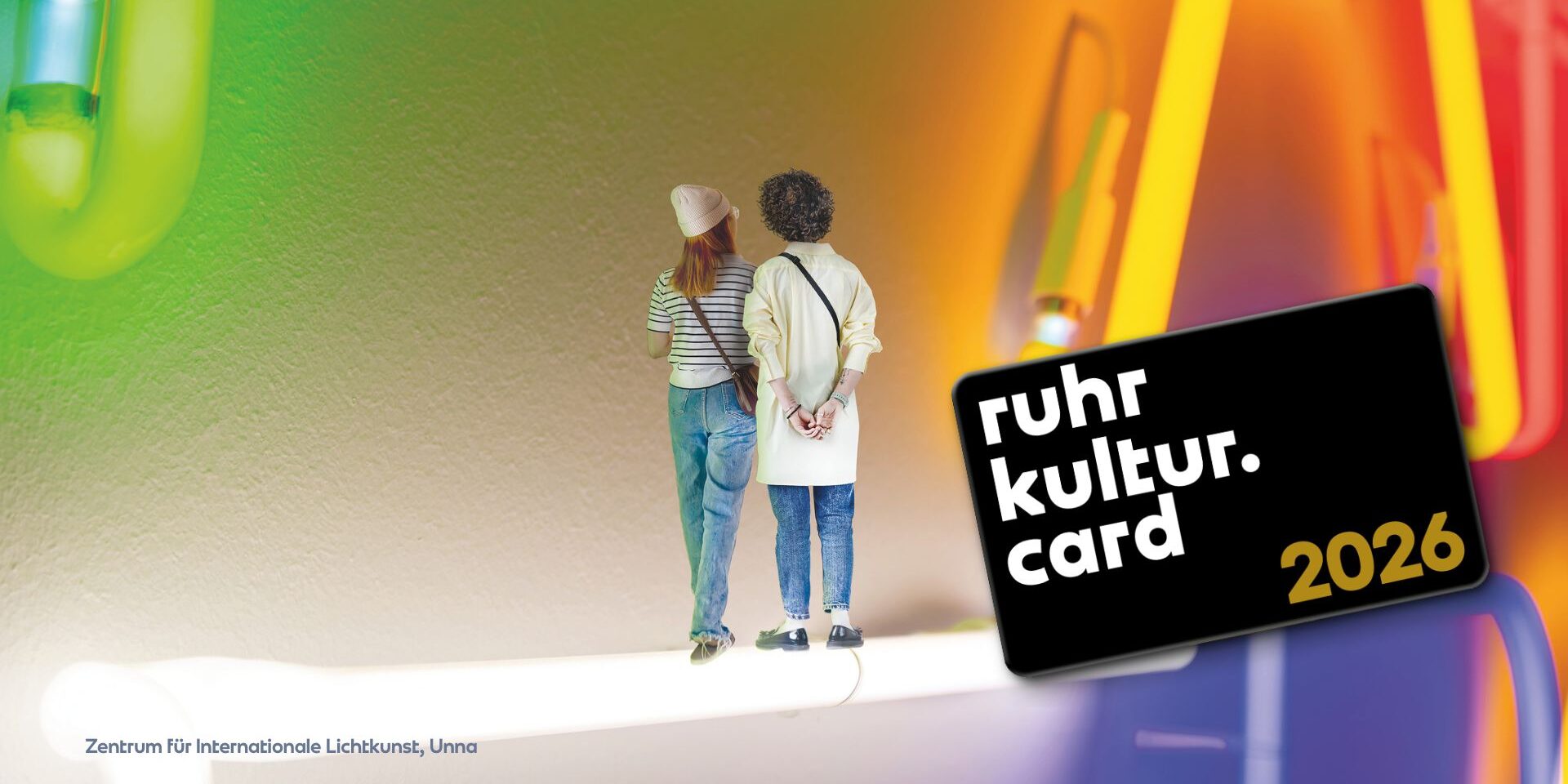 Two people stand side by side on a brightly lit surface, facing a colorful, abstract neon background. The text “ruhr kultur. card 2026” appears on a black card in the foreground.