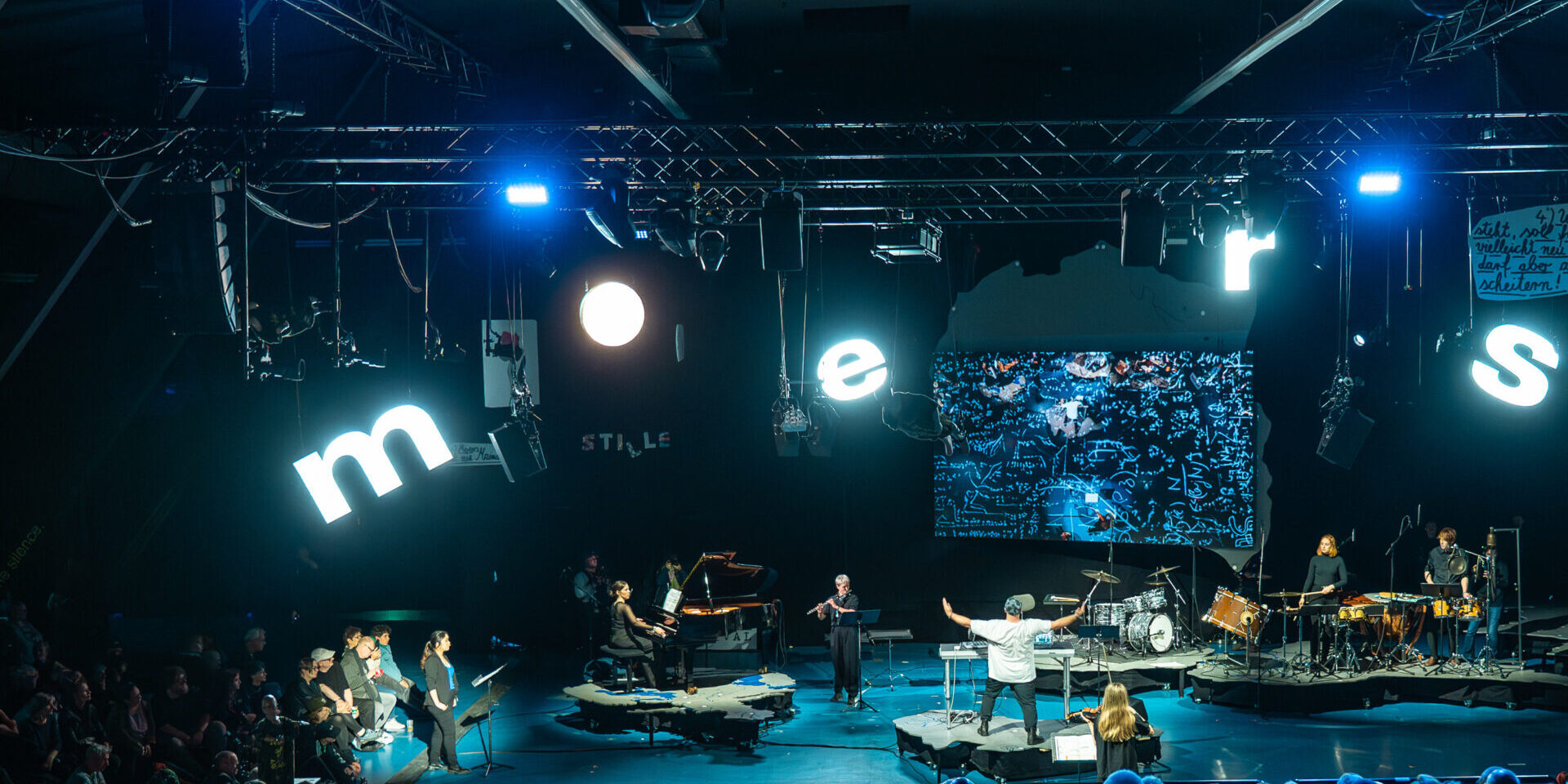 A large audience watches a live music performance on a stage with blue lighting. The word moers is spelled out in large illuminated letters above the stage. Several musicians and instruments are visible.
