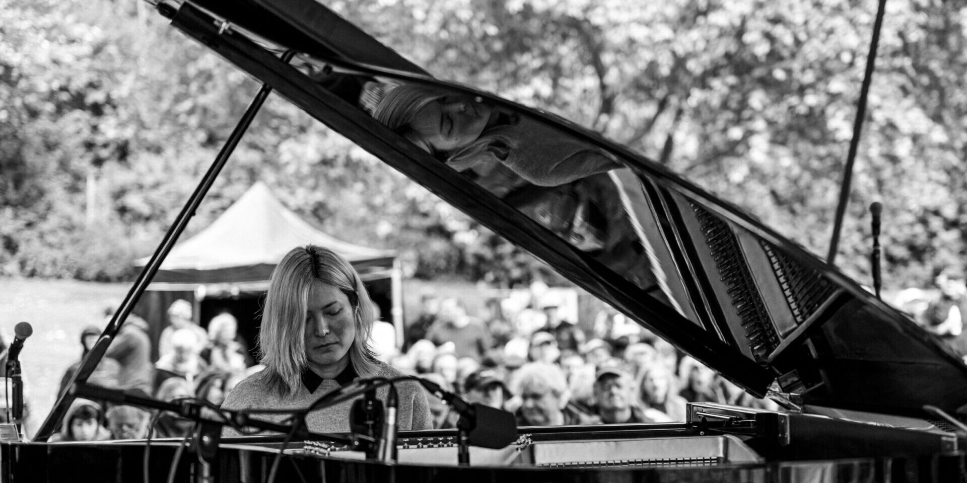 A woman plays a grand piano outdoors, her reflection visible on the open lid. A crowd watches in the background, and trees fill the scene. The image is in black and white.