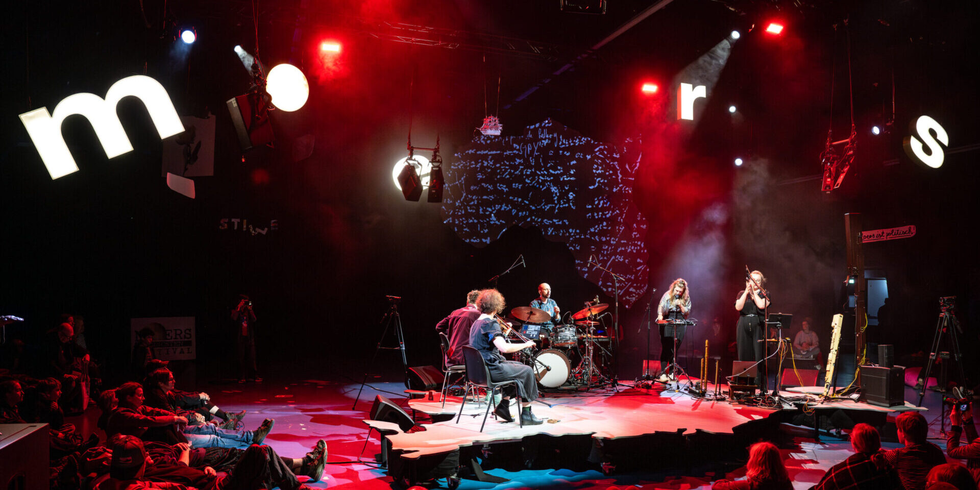 A band performs on a stage shaped like a map, surrounded by seated and standing audience members. Large illuminated letters hang above, and red and purple lighting creates a dramatic atmosphere.