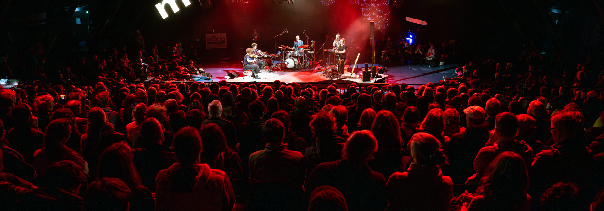 A large audience watches a concert on a dimly lit stage with red lights. The word moers is displayed in large letters above the stage, and a band performs under the lights.