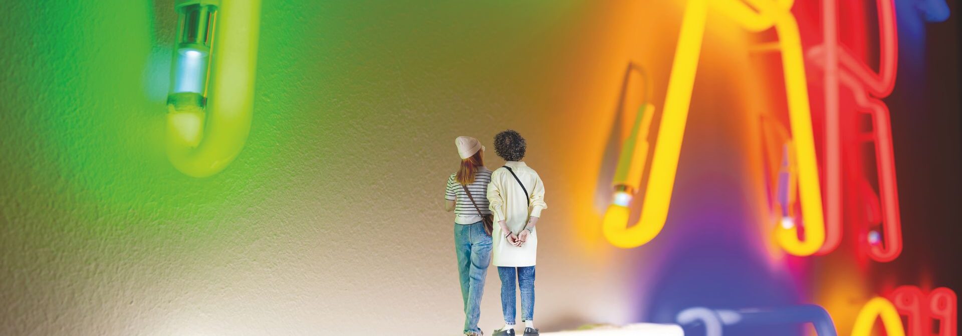 Two small, digitally shrunken people stand on a glowing white neon tube, facing a colorful, oversized neon sign on a wall. The sign features large red, yellow, blue, and green illuminated letters.