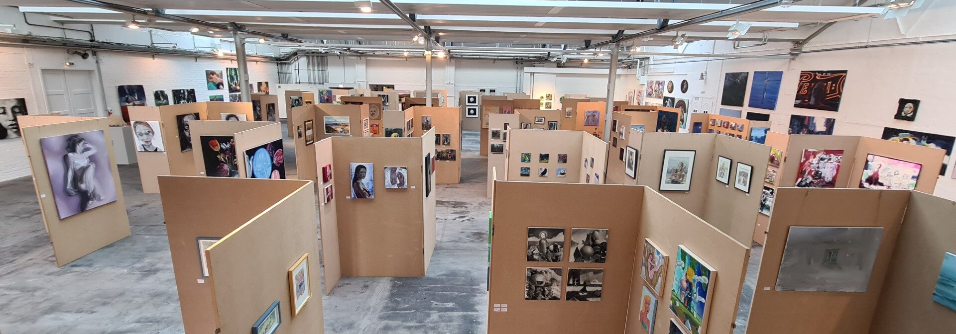 An art gallery with numerous beige display panels arranged in rows, each showcasing various paintings and photographs. The space is well-lit with natural and artificial light, and artwork covers most of the panels.