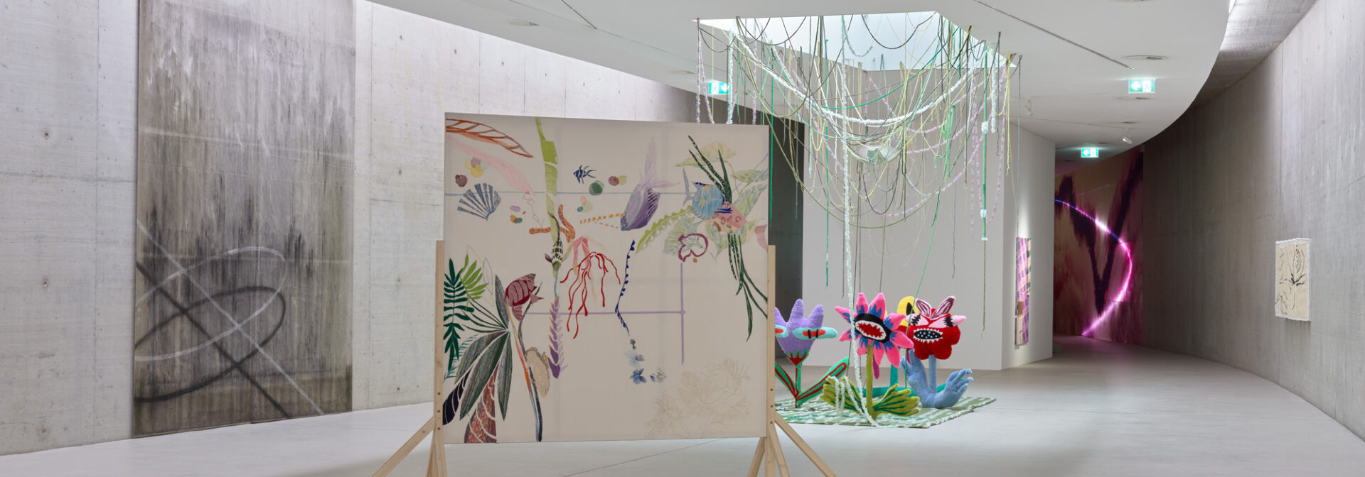 A modern art gallery with minimalist concrete walls displays colorful artwork, including a floral painting on wheels, vibrant flower sculptures, and hanging green light strings, in a bright, curved hallway.