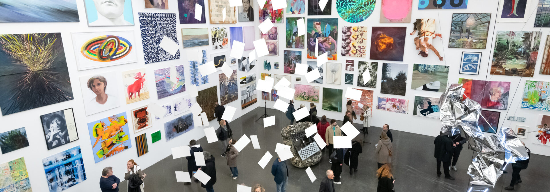 A group of people view artwork in a brightly lit gallery with colorful paintings covering the walls and white papers or cards suspended from the ceiling as part of the exhibit.
