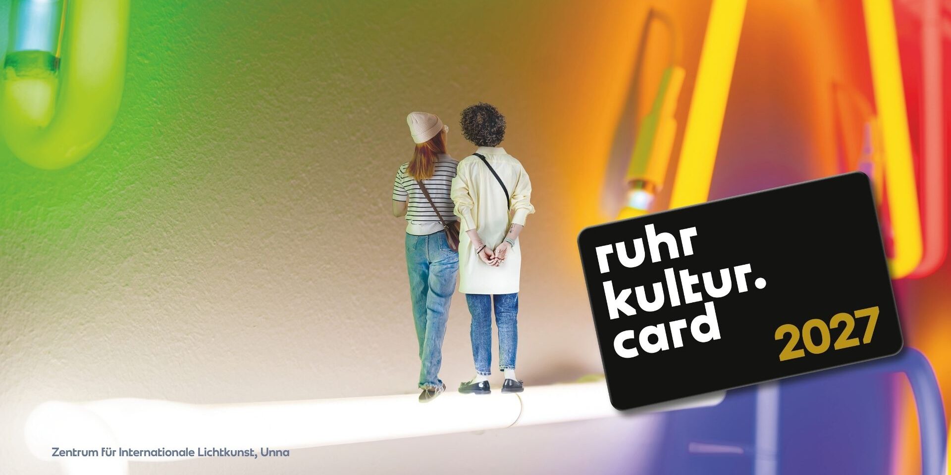 Two people stand side by side on a brightly lit surface, facing a colorful, abstract neon background. The text “ruhr kultur. card 2027” appears on a black card in the foreground.